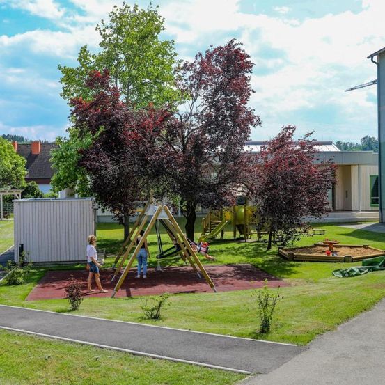 Spielplatz im Freien mit rotem Gummiboden, Bäumen und Rutschen. Eine Frau geht und ein Mädchen rutscht herunter.