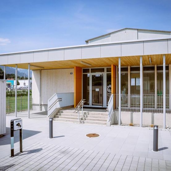 Ein modernes weißes Gebäude mit orangefarbenen Akzenten und Glastüren und -fenstern. Treppen führen zum Eingang, mit Metallgeländern auf beiden Seiten.