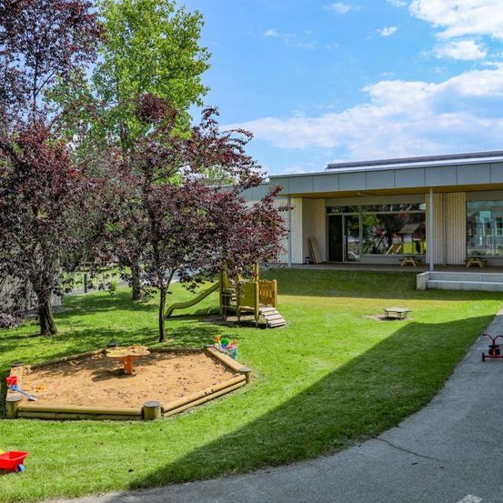Ein Spielplatz im Freien verfügt über eine Rutsche, einen Sandkasten und eine Bank im Schatten von Bäumen. Das Gebäude hat Glasfenster und eine Veranda.