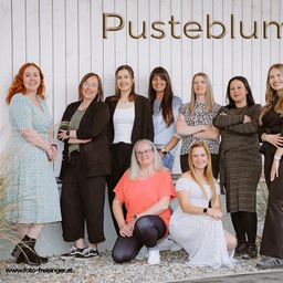 Eine Gruppe von Frauen in verschiedenen Outfits posiert für ein Foto vor einer weißen Wand mit der Aufschrift 'Pusteblum'.