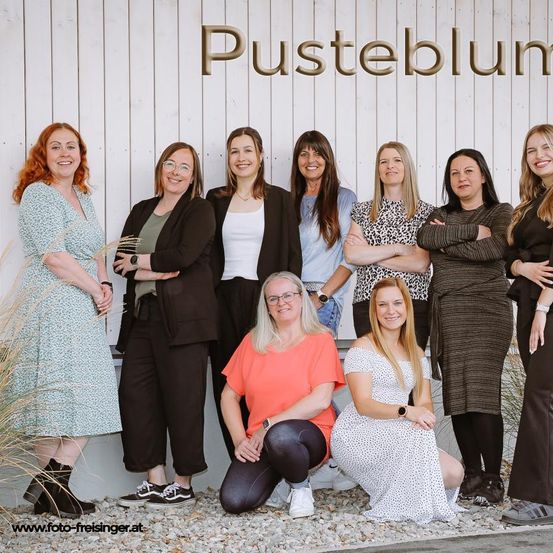 Eine Gruppe von Frauen in verschiedenen Outfits posiert für ein Foto vor einer weißen Wand mit der Aufschrift 'Pusteblum'.