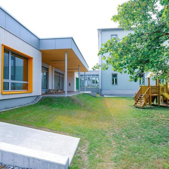 Ein modernes Gebäude mit gelben Akzenten und Glasfenstern. Es gibt Tische im Freien und einen Spielplatz.