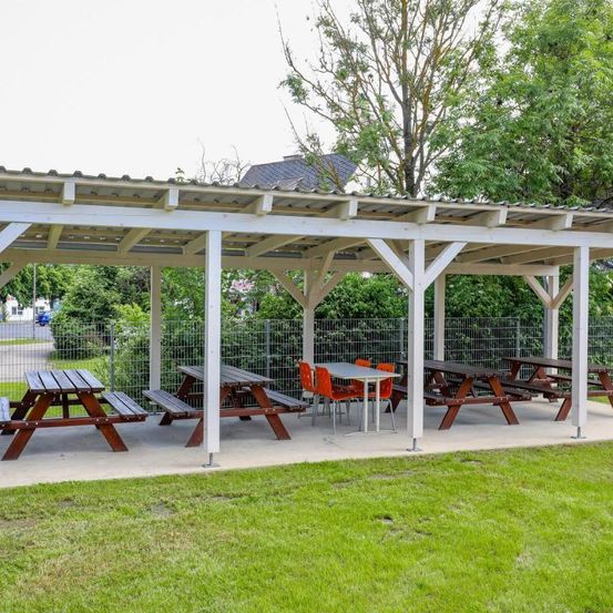 Ein Freiluft-Picknickplatz mit Holztischen und Stühlen unter einem weißen Pavillon, umgeben von Grün und einem Zaun. Der Bereich ist leer.
