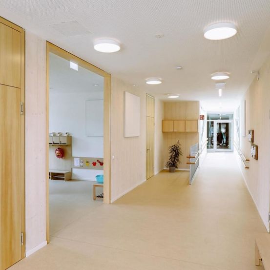 Ein leerer Flur mit weißer Wand, beigem Boden und Holztüren. In der Nähe der Tür stehen ein Topf mit Pflanzen und ein kleiner Tisch.