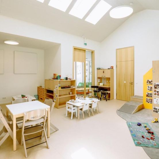 Ein Raum mit weißen Wänden, einer weißen Decke und weißen Tischen. Holzmöbel sind im Raum aufgestellt. Der Raum hat einen Spielbereich mit Spielzeug.