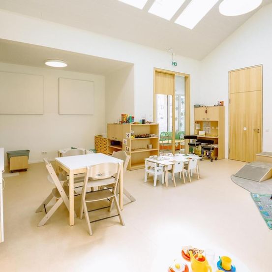 Ein Kinderzimmer mit weißen Wänden, einem weißen Tisch und Stühlen sowie Regalen mit Spielzeug und Büchern. Es gibt eine Holztür, einen Mülleimer und einen Teppich.