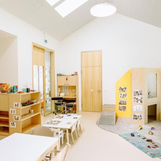 Ein sauberer und geräumiger Klassenraum mit weißen Wänden, Holzmöbeln, einem weißen Tisch, Stühlen und einer gelben Wand mit Spielbereich und Spielzeug.