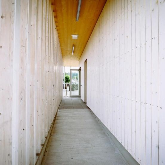 Ein leerer Korridor mit hölzernen Wänden und Decke, der zu einer Tür mit einem Metalltor führt, beleuchtet von Deckenleuchten.