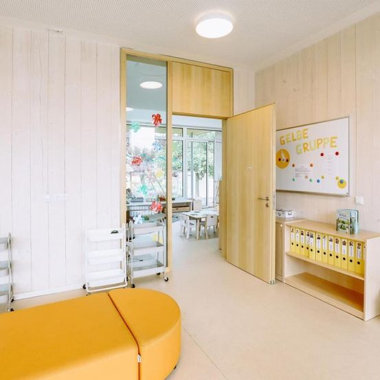 Ein heller Raum mit Holzwänden, einem gelben Sofa und einem Regal mit gelben Ordnern. Eine Tür führt in einen anderen Raum mit Tisch und Stühlen. Eine Tafel mit 'GELBE GRUPPE' ist an der Wand.