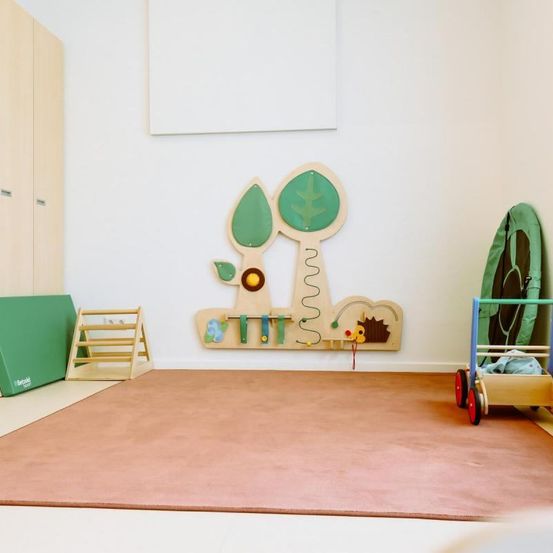 Ein Spielzimmer mit Holzmöbeln und Teppichboden, das eine Wand mit Baumdesign und einer Rutsche aufweist. Ein Holzwagen mit grüner Abdeckung und eine Holztreppe sind ebenfalls vorhanden.