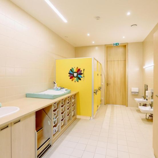 Ein sauberer und geräumiger Wickelraum mit gelben Wänden und einem weißen Fliesenboden, ausgestattet mit einer Wickelkommode, Schränken und Waschbecken.