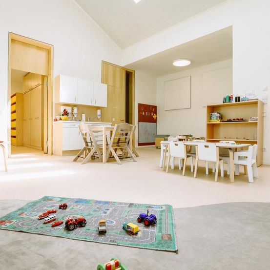 Ein Kinderspielzimmer mit Teppich, Spielzeugautos und Tischen. Es hat Holzmöbel, weiße Wände und eine Deckenleuchte.