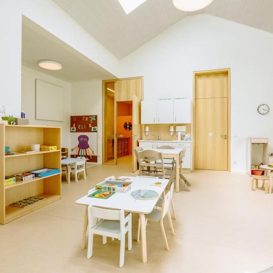 Ein Raum mit weißen Wänden und einem Holzboden. Es verfügt über einen Esstisch mit Stühlen, ein Holzregal mit Büchern und eine Küche mit Arbeitsplatte und Schränken.