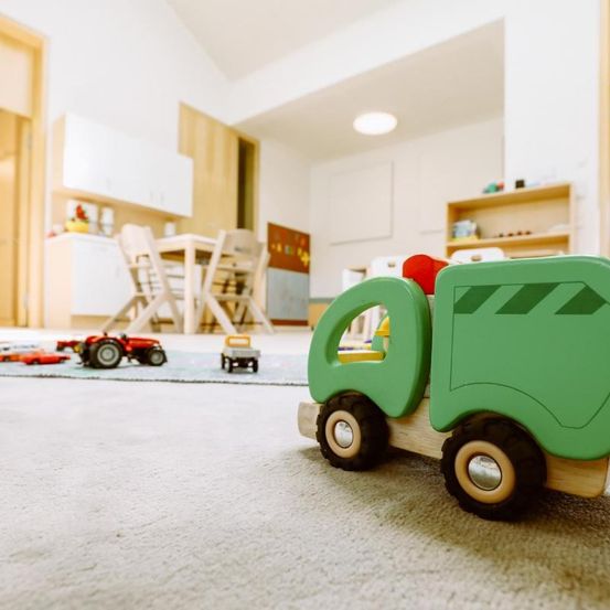 Ein Spielzeuglastwagen aus Holz steht auf einem Teppich in einem Spielzimmer. Im Hintergrund befindet sich ein Essbereich mit Stühlen und einem Tisch.