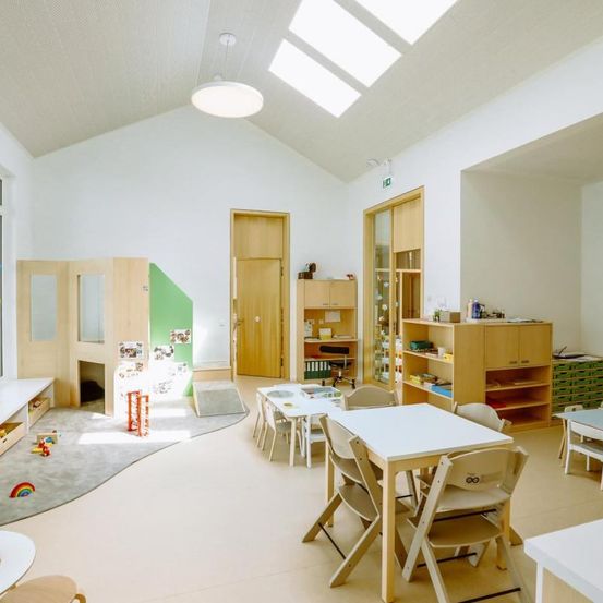 Ein heller, luftiger Raum mit weißen Wänden und Holzmöbeln. Ein großer Tisch mit Stühlen steht in der Mitte. Ein Regal mit verschiedenen Gegenständen befindet sich rechts und ein kleiner Teppich liegt auf dem Boden.