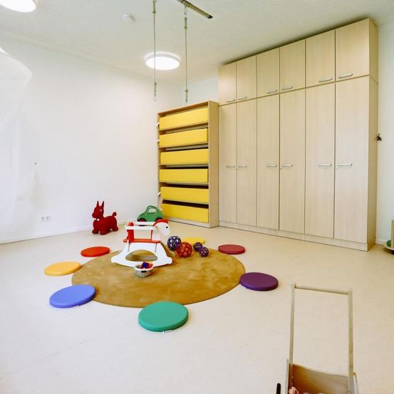 Ein Spielzimmer mit einer weißen Wand, einem braunen Boden und einem runden Teppich mit Spielzeug und Bällen. Holzschränke und Regale befinden sich an der Seite.