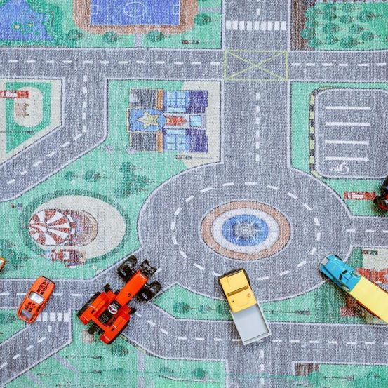 Ein Spielmatte mit Straße und Autos. Es gibt Gebäude und einen Spielplatz. Die Matte hat ein Fußballfeld und einen Parkplatz.