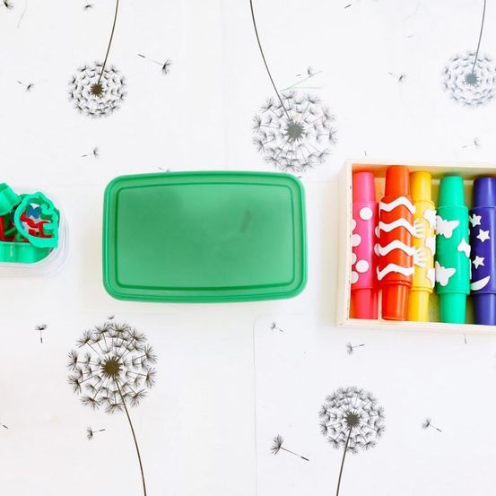Ein weißer Hintergrund mit Löwenzahn, einer grünen Box mit Deckel, einer Box mit Buntstiften und einer Box mit Ausstechern.