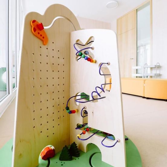 Eine Holzspielstruktur mit verschiedenen farbigen Perlen und einer orangefarbenen Fischform. Die Struktur steht auf einer grünen Basis in einem sauberen, modernen Raum.