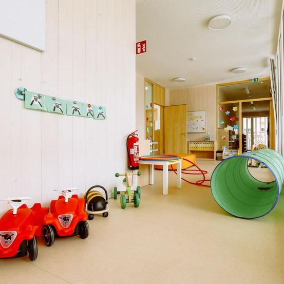 Ein Spielbereich für Kinder mit Spielzeug und einem Feuerlöscher. Es gibt einen Tunnel, einen Tisch und einen roten Schlauch auf dem Boden.