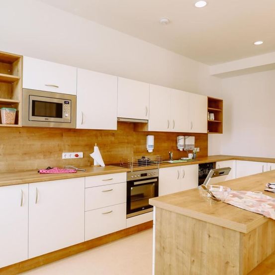 Eine moderne Küche mit weißen Schränken und Holzarbeitsplatten. Ein Ofen und eine Mikrowelle sind an der Wand installiert. Eine Spüle mit Wasserhahn befindet sich in der Ecke.