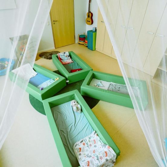 Fünf grüne Kinderbetten sind in einem Kreuz angeordnet auf einem Boden mit weißen und gelben Fliesen. Sie sind mit Stoff bedeckt und einige sind mit Spielzeug ausgestattet. Ein weißer Baldachin hängt über sie.