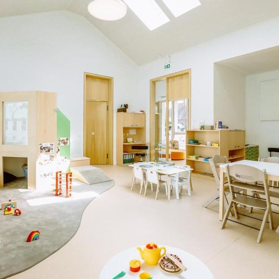 Ein heller Kinderzimmer mit weißen Wänden, Holztüren und einem Spielbereich mit Spielzeug. Es gibt Tische und Stühle zum Sitzen, Regale mit Büchern und einen kleinen Schreibtisch.