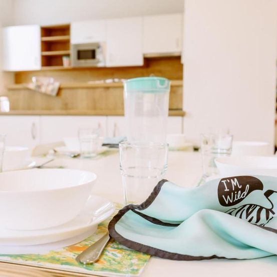 Ein Küchentisch ist mit Tellern, Gläsern und einem Krug gedeckt. Eine weiße Schüssel steht auf einer Untertasse. Ein Serviettentuch mit dem Aufdruck 'I'm Wild' liegt auf dem Tisch.