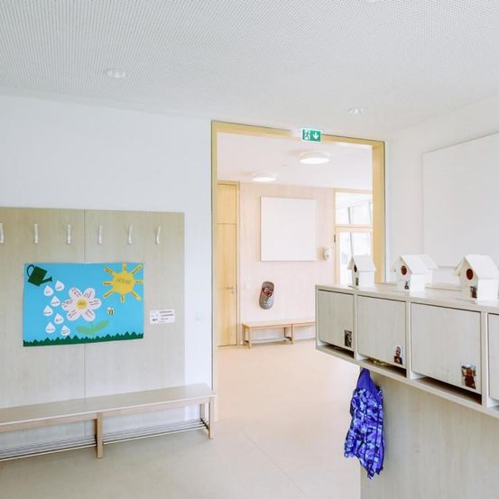 Ein sauberer, heller Raum mit weißen Wänden, einer Holzbank und einer Tafel mit einer bunten Zeichnung einer Blume und einer Sonne.