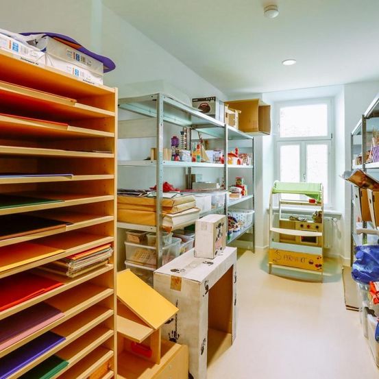 Ein Raum gefüllt mit Regalen und Lagerboxen, ein Schreibtisch davor, ein Spielzeugküchenset und ein Fenster mit Blick nach draußen.