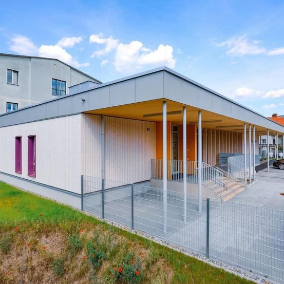 Ein modernes Gebäude mit grauer Außenfassade, lila Türen und einem gelben Dach. Es hat einen eingezäunten Eingang mit Treppen. Ein Auto ist in der Nähe geparkt.