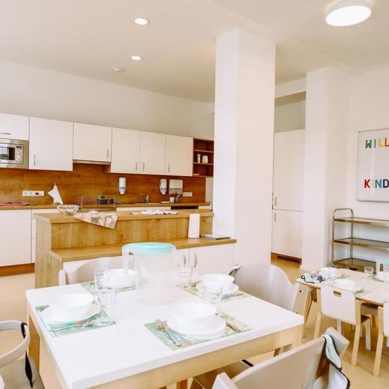 Eine moderne Küche mit weißen Schränken, einer Holzarbeitsplatte, einem Kühlschrank und einem Essbereich mit Stühlen und einem Tisch. Der Tisch ist mit Tellern, Schüsseln, Gläsern und einer Kanne gedeckt.