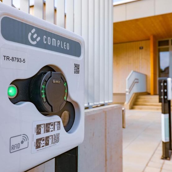 Nahaufnahme einer weißen Ladestation für Elektrofahrzeuge mit der Aufschrift COMPLEO und einer grünen Kontrollleuchte. Die Station steht vor einem Gebäude mit einer Treppe.
