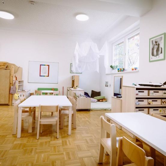 Ein aufgeräumtes Klassenzimmer mit Holzmöbeln. Eine Tafel und Bilderrahmen schmücken die weißen Wände. Der Raum verfügt über einen kleinen Tisch und Stühle, einen Schrank mit Schubladen und ein Fenster mit Blick auf Bäume.