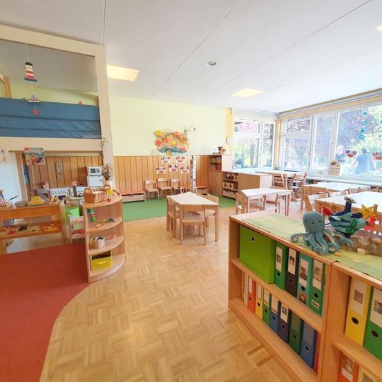 Ein Innenklassenraum mit Holzboden, Tischen, Stühlen, Regalen mit Büchern, Spielzeug und einem Spielbereich mit Teppich. Es gibt Fenster mit Blick nach draußen.