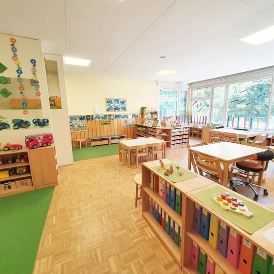 Ein Kinderklassenzimmer mit Holztischen und -stühlen, Regalen mit Büchern und einem grünen Boden. Es gibt bunte Spielzeuge auf einem Regal und Kunst an den Wänden.