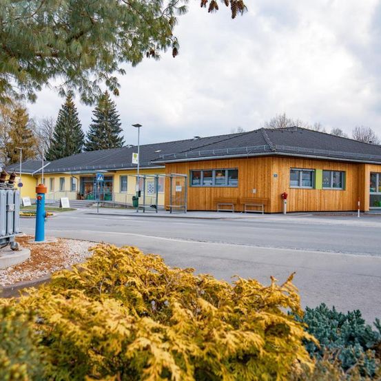 Ein gelbes Gebäude mit grauem Dach steht an einer Straße, umgeben von Grün. Bänke und ein Feuerlöscher stehen davor. Bäume und ein bewölkter Himmel im Hintergrund.
