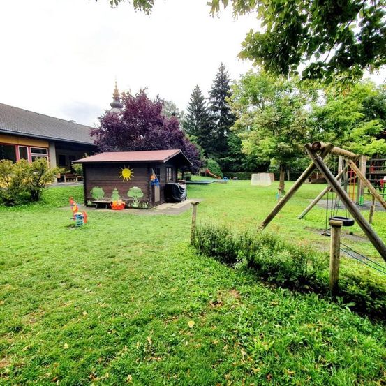 Ein Spielplatz mit einem kleinen Holzhaus, einer Schaukel und Rutschen. Bäume und Pflanzen umgeben das Gebiet, mit einem Gebäude im Hintergrund.