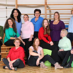 Eine Gruppe von Frauen in verschieden farbigen Oberteilen und Hosen lächelt und posiert für ein Foto in einem Raum mit Holzboden und Kletterseilen im Hintergrund.