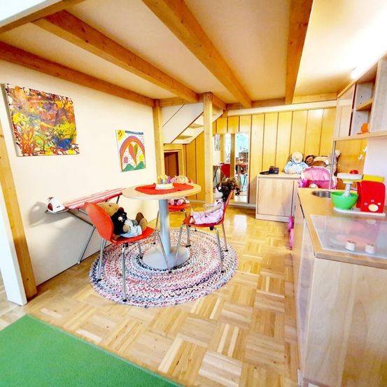 Ein gemütlicher Essbereich mit rundem Tisch und Stühlen, bunte Kunstwerke an der Wand, eine Küchenzeile mit Waschbecken und Holzschränken. Es gibt Spielzeug und einen grünen Teppich auf dem Holzboden.