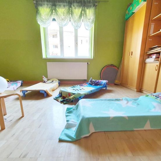Ein Kinderzimmer mit Holzböden und grünen Wänden. Drei kleine Betten mit sternförmigen Decken liegen auf dem Boden. Ein Holzstuhl und ein Schrank sind im Zimmer. Ein weißer Heizkörper ist an der Wand montiert.