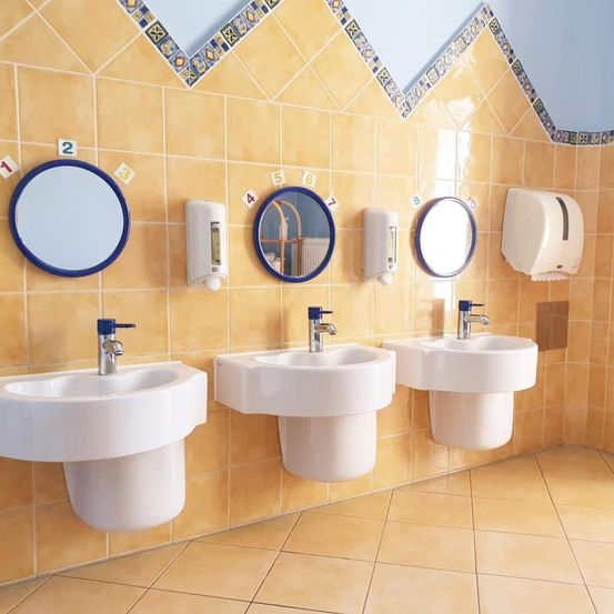 Ein Badezimmer mit drei Waschbecken und drei Spiegeln an gelben gefliesten Wänden. Über den Waschbecken befinden sich Seifenspender und über den Spiegeln nummerierte Schilder.