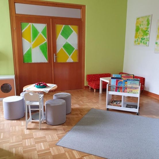 Ein Raum mit einer Holztür mit farbenfrohen Glasscheiben. Im Inneren befindet sich ein Tisch mit Stühlen, ein Bücherregal mit Büchern und eine gemütliche Couch. Die Wände sind grün und weiß gestrichen, und es gibt einen Teppich auf dem Holzboden.