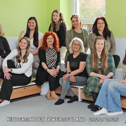 Eine Gruppe von Frauen, lächelnd und sitzend, in verschiedenen Kleidungsstücken, vor einer grünen Wand mit Fenster. Der Text 'KINDERGARTEN ZWERGENLAND 2024/2025' befindet sich unten.