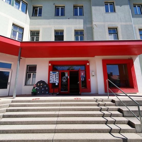 Der vordere Eingang eines Kindergartens mit rotem Rand. Ein Schild mit der Aufschrift 'Kindergarten' befindet sich über dem Eingang. Treppen führen mit einer Handlauf zum Eingang.
