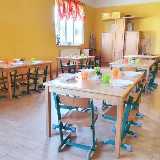 Ein Klassenzimmer mit gelben Wänden und Holztischen und -stühlen. Jeder Tisch hat einen Teller, einen Löffel und eine Topfpflanze. In der Ferne befinden sich weitere Tische und Stühle.