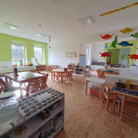 Ein hell beleuchteter Klassenraum mit Holzmöbeln, grünen Wänden und bunten Dinosaurier-Spielzeug, die von der Decke hängen. Es gibt Tische, Stühle und Regale voller Gegenstände.