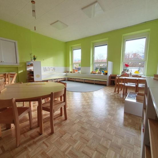 Ein heller Raum mit grünen Wänden, Holzboden und mehreren Tischen und Stühlen. Der Raum verfügt über einen runden Tisch mit Stühlen, einem Regal mit Pflanzen und Fenstern mit Blick nach draußen.