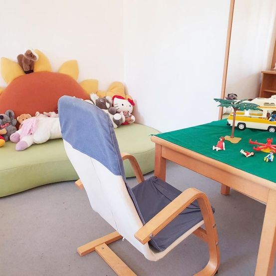 Ein Kinderspielzimmer mit einem mit Spielzeug bedeckten Tisch und einem gepolsterten Stuhl. Gefüllte Plüschtiere sind auf der Couch. Die Wand ist weiß gestrichen.
