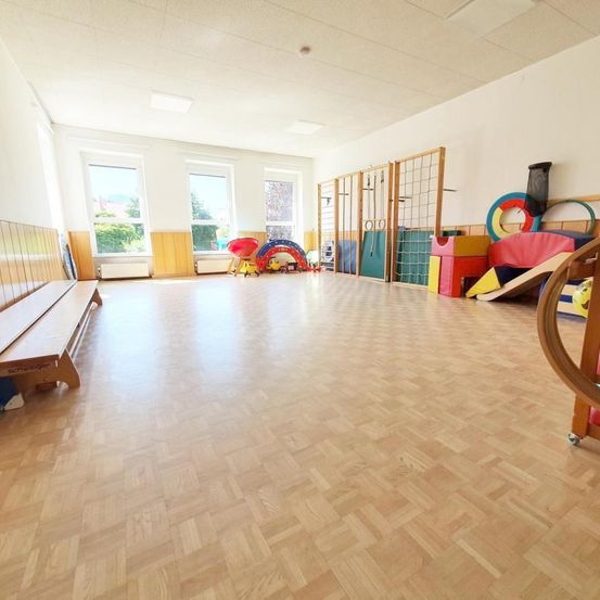 Ein leerer Raum mit Holzböden und einem großen offenen Bereich. Verschiedenes Spielzeug und Spielgeräte sind auf der rechten Seite verstreut. Es gibt zwei Fenster an der linken Wand und eine Bank auf der linken Seite.